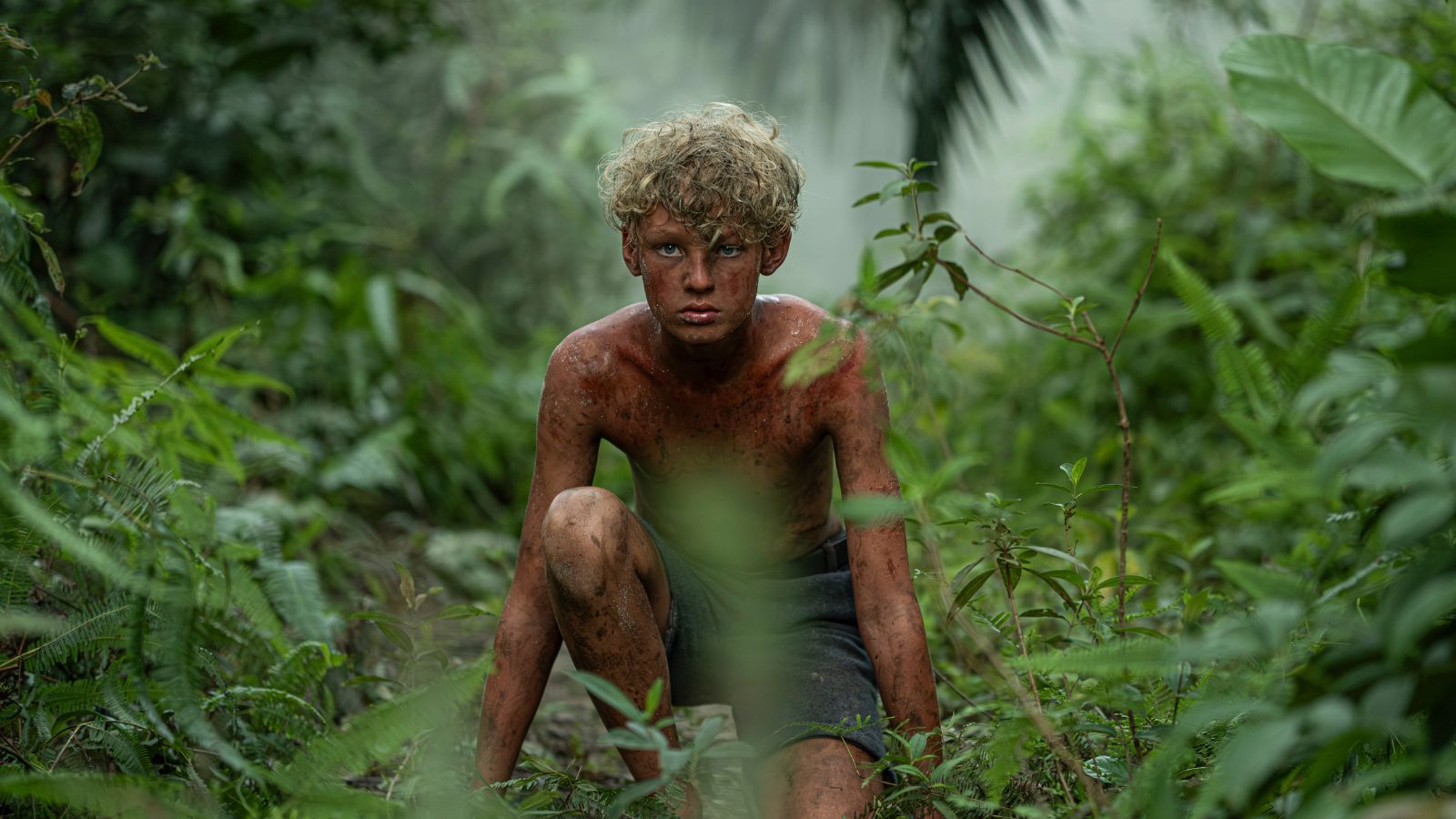 A person with curly blond hair sits on the ground in a dense jungle environment. The individual appears muddy and scratched, surrounded by lush green foliage and mist, evoking a wild and untamed setting.