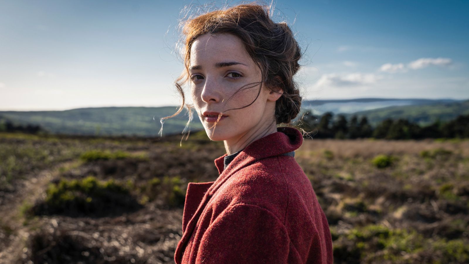 A person in a red coat stands in an open countryside landscape with rolling hills and a clear blue sky. The ground is covered in grass and shrubs, and the scene conveys a sense of wide, natural space.