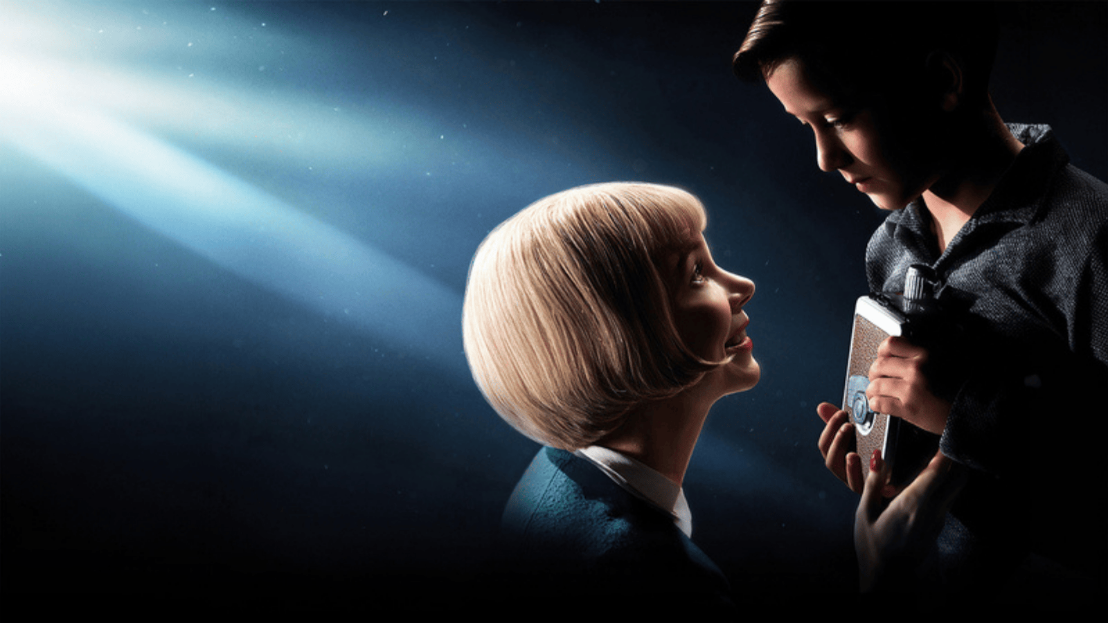 Two people standing together in dramatic spotlighting, with one holding a vintage film camera against a dark, cinematic background.