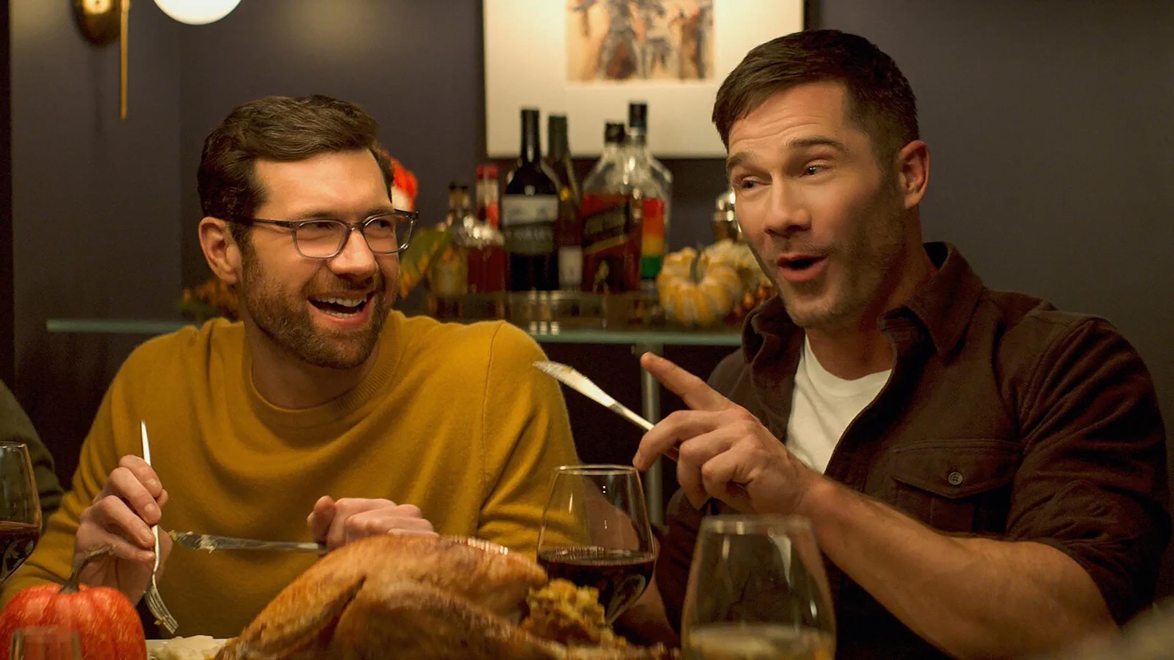 A man with a yellow humper and glasses eating a thankgiving meal sat next to a man in a brown shirt holding a knife in his left hand