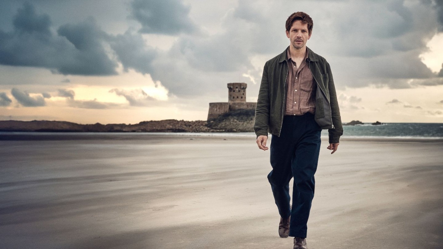Promotional image for the TV series Bergerac. Damien Molony as Jim Bergerac walks towards the camera across a wide, flat beach under a moody, overcast sky. He is dressed in an open dark olive jacket, a brown shirt, and navy trousers. In the background, a historic stone coastal tower stands on a rocky outcrop against the sea