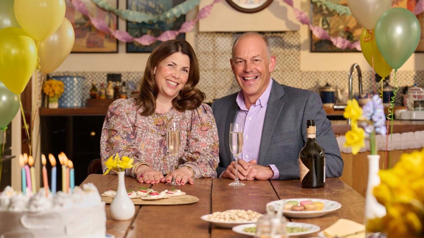A women in a flowery dress and a man in a suite sat at a table with balloons and cake