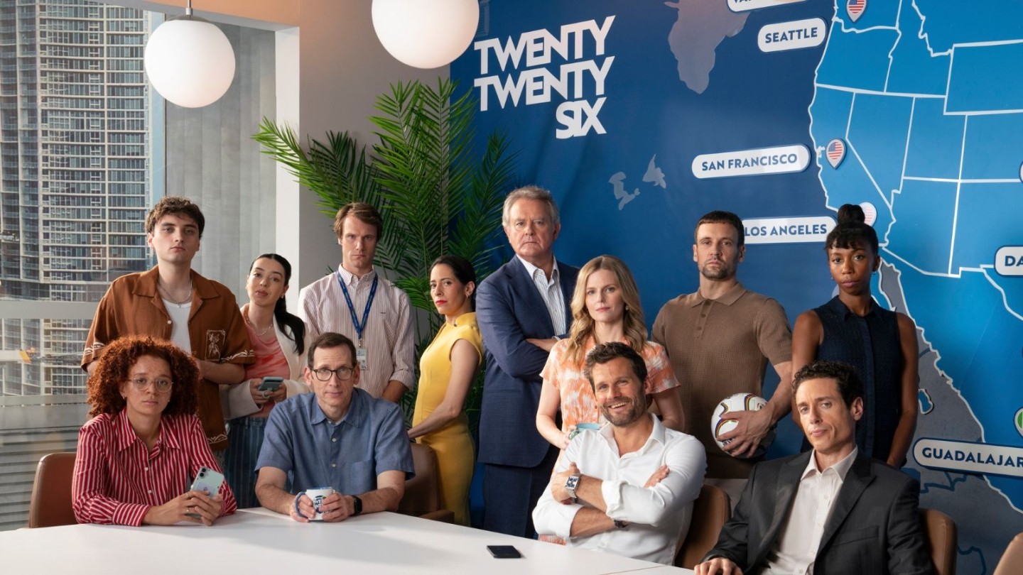 A group of people gathered around a modern conference table in a bright office. Behind them is a large blue wall map with city names like Seattle, San Francisco, Los Angeles, and Guadalajara, along with the text “TWENTY TWENTY SIX.” Two spherical pendant lights hang from the ceiling.
