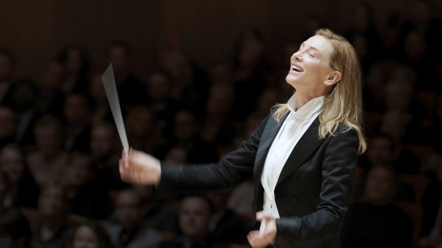 Orchestral conductor wearing a formal black tailcoat and white dress shirt, holding a baton mid-performance against a dark concert hall background.
