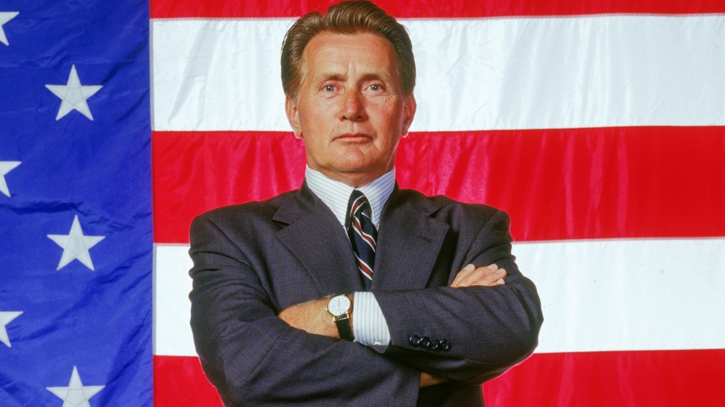 A man in a dark business suit stands confidently with his arms folded in front of a large American flag, creating a strong, authoritative and patriotic image.