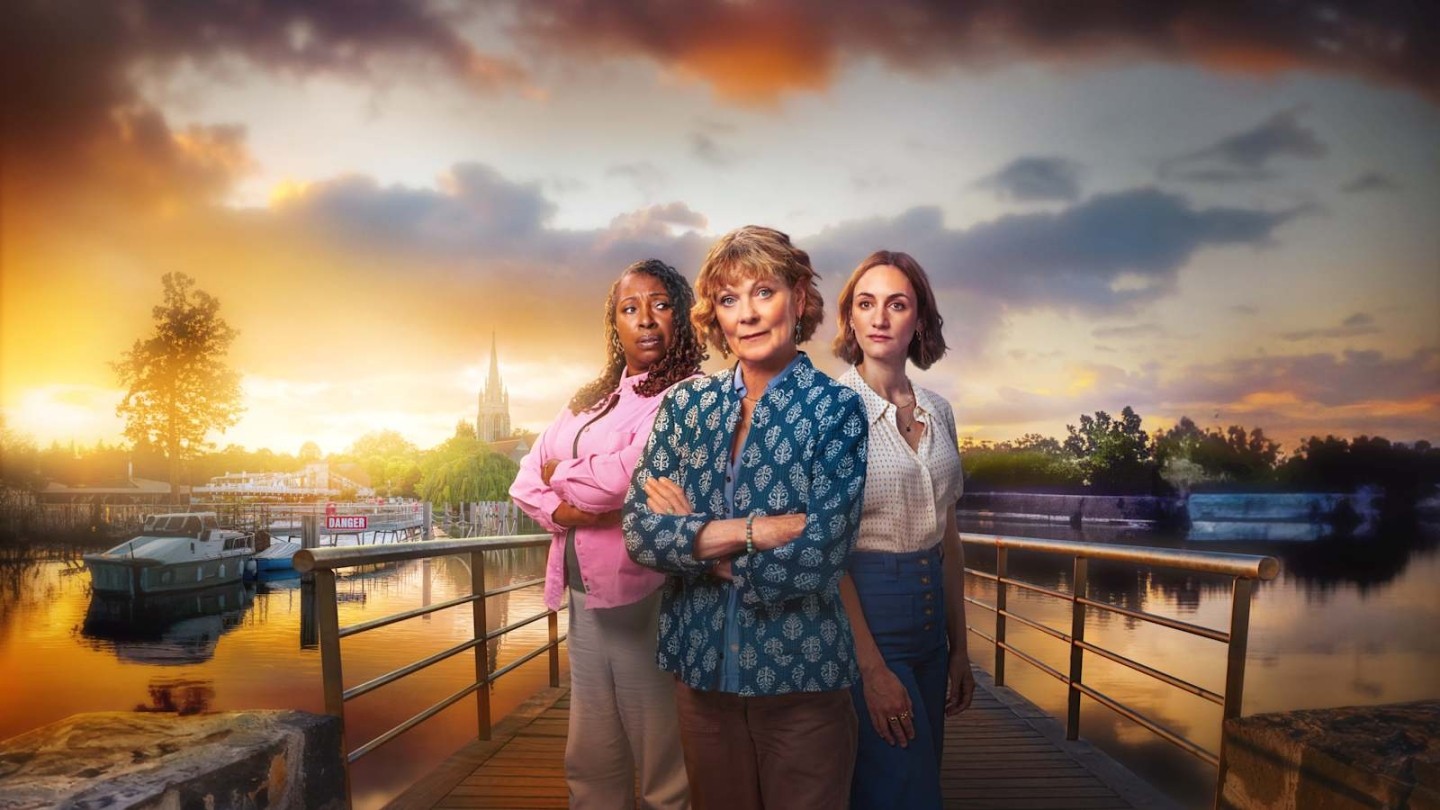 Three people stand confidently on a wooden bridge overlooking a calm riverside at sunset. Warm golden light reflects off the water, highlighting boats moored along the banks and a distant church spire rising above the trees. The trio is positioned at the centre of the bridge, dressed in casual but stylish clothing, with a dramatic, colourful sky creating a serene yet cinematic atmosphere