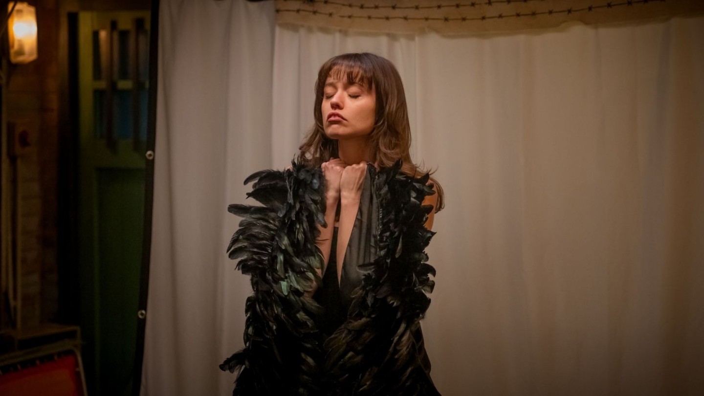 Person dressed in a feathered costume performing on a stage with warm lighting and a curtain backdrop.