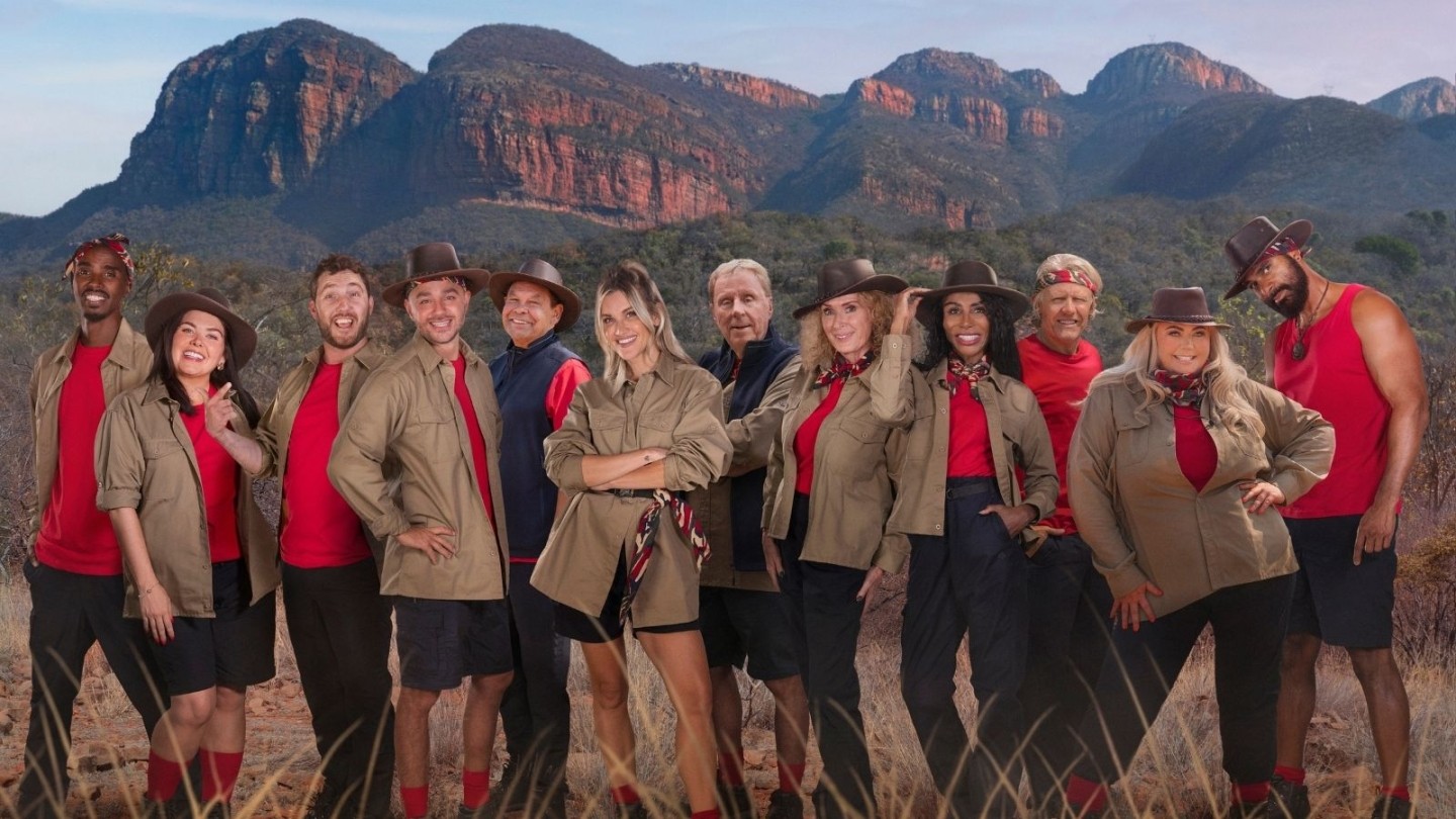 Group of I’m a Celebrity South Africa cast members posing together in front of a rugged mountain backdrop, wearing red tops and khaki uniforms.