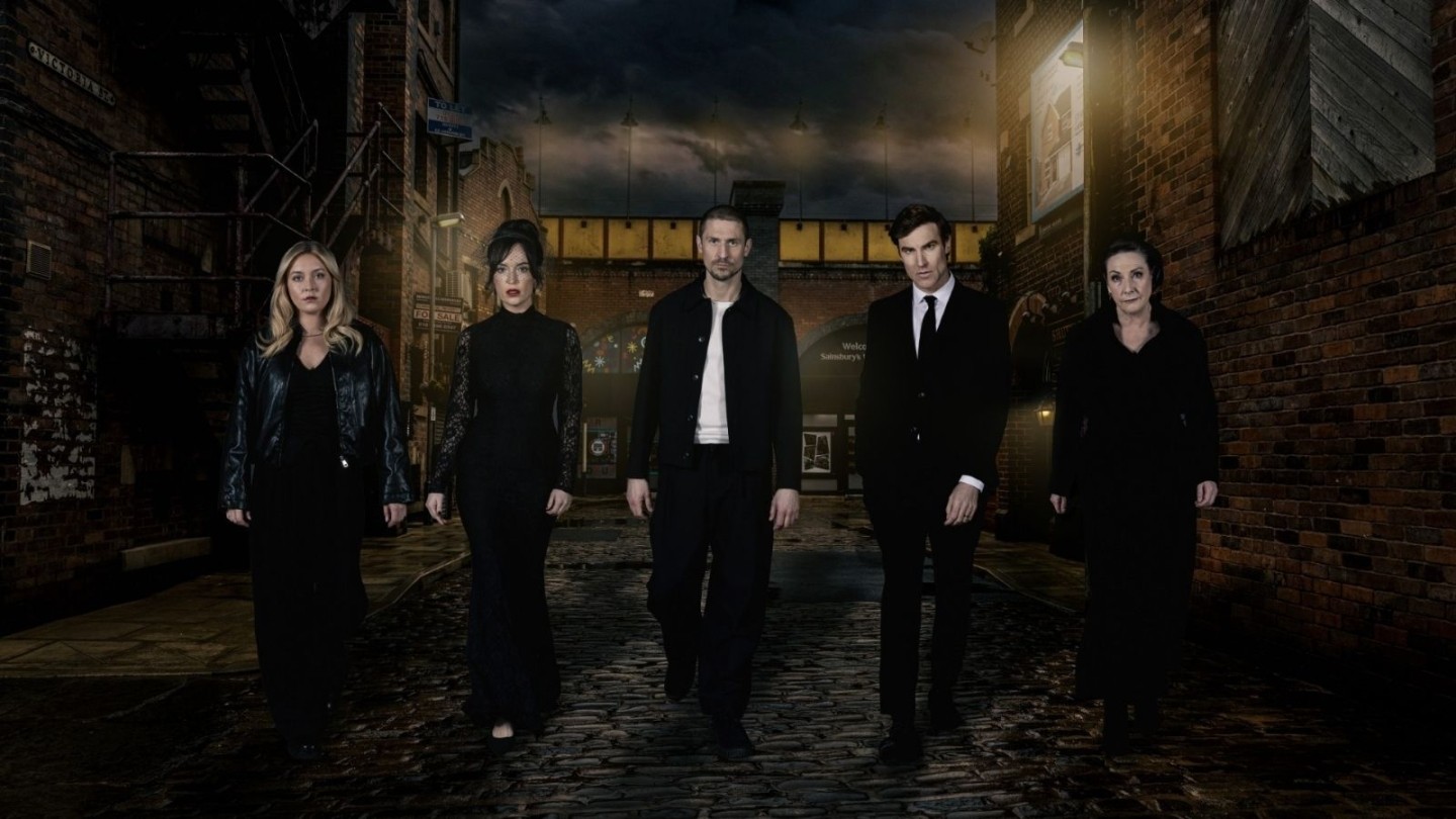 Six people walk towards the camera along a dark, cobbled street at night, surrounded by brick buildings and streetlights, with a railway bridge behind them.