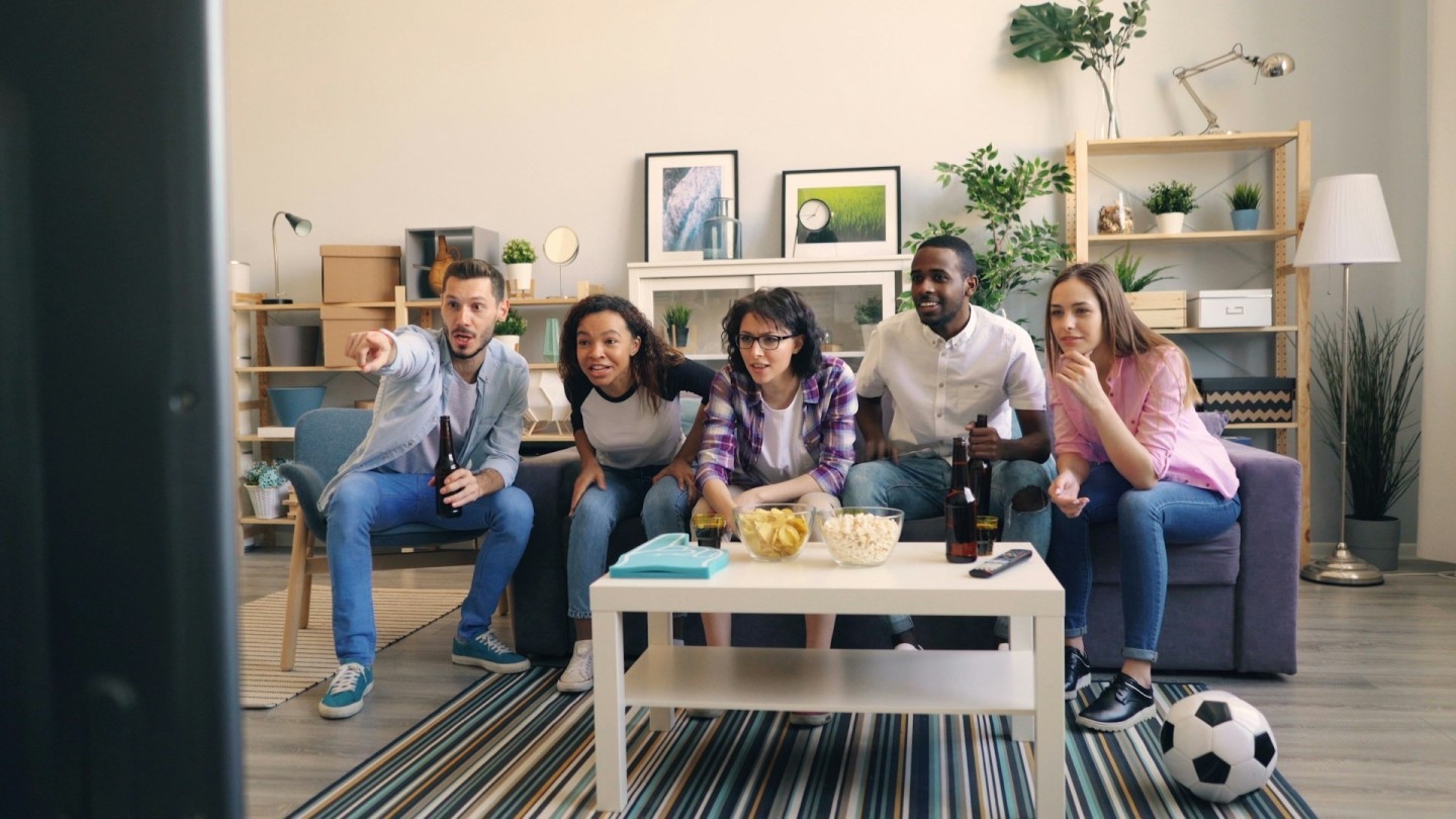 Five football fans sit on the sofa watching and reacting to a match on their TV