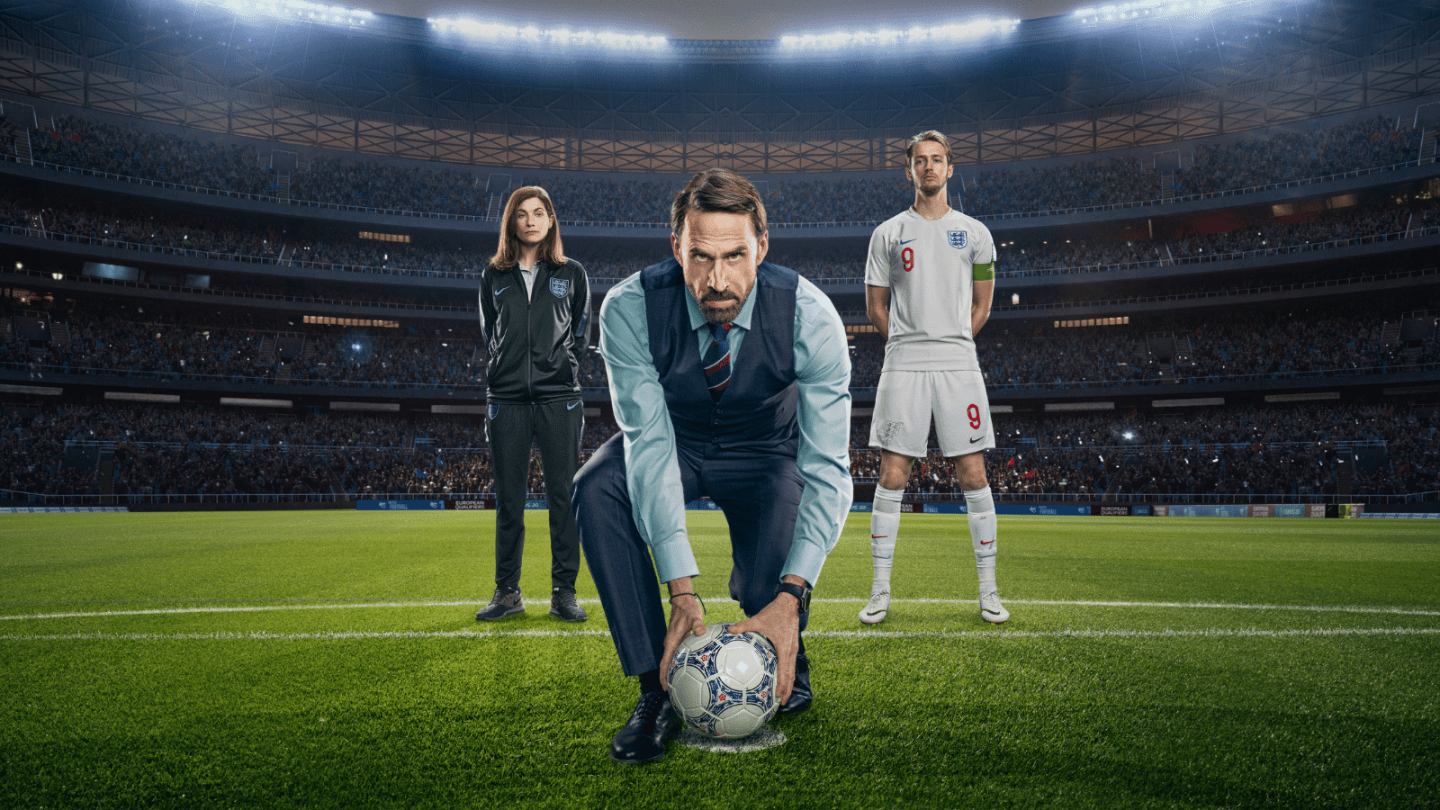 Joseph Fiennes stood in a smart outfit holding a football on a football pitch with a female coach behind him to the left and an England football player to the right