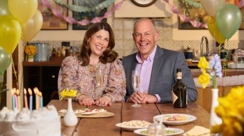 A women in a flowery dress and a man in a suite sat at a table with balloons and cake
