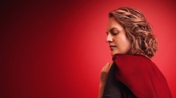 Elizsbeth Moss as June in a Handmaids tale, wearing a red cape against a red black ground