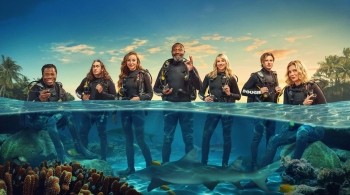 4 men and 3 women dressed in scuber gear sat on rocks in a the sea