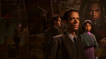 Three people dressed in vintage-style clothing stand in a dimly lit room with walls covered in old theatrical posters featuring words like “Vampire” and “Satan.” The figures wear formal suits and period dresses, evoking a dark, gothic atmosphere.