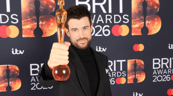 A person in a black outfit holds up a BRIT Awards 2026 trophy in front of a backdrop featuring BRIT Awards logos, sponsor icons and the ITV logo.
