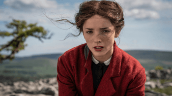 A person wearing a red coat sits outdoors on a rocky landscape with rolling hills and a partly cloudy sky in the background. Loose strands of hair blow in the wind. 