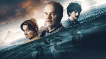 Three people stand waistdeep in dark water, their reflections visible beneath the surface. A dramatic sky and distant rocky coastline create an eerie, atmospheric backdrop. 