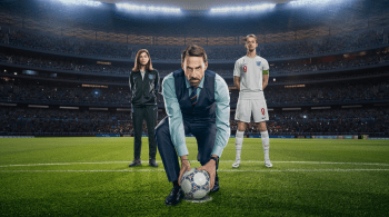 Joseph Fiennes stood in a smart outfit holding a football on a football pitch with a female coach behind him to the left and an England football player to the right