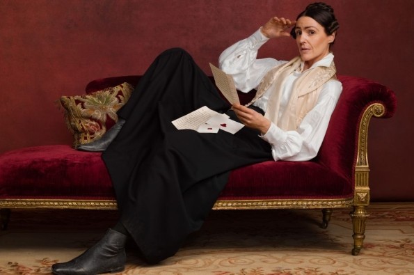 Promotional image for the TV series Gentleman Jack. Suranne Jones as Anne Lister is reclining on a luxurious red velvet chaise longue against a dark red wall.