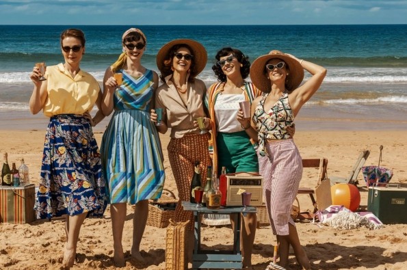 The cast of Ten Pound Poms stood on a beach in 50s clothing