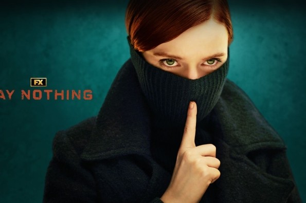 A women with aurburn hair and green eyes stood with half her face covered behind a scarf and her finger against where her lips would be, staring int the camera