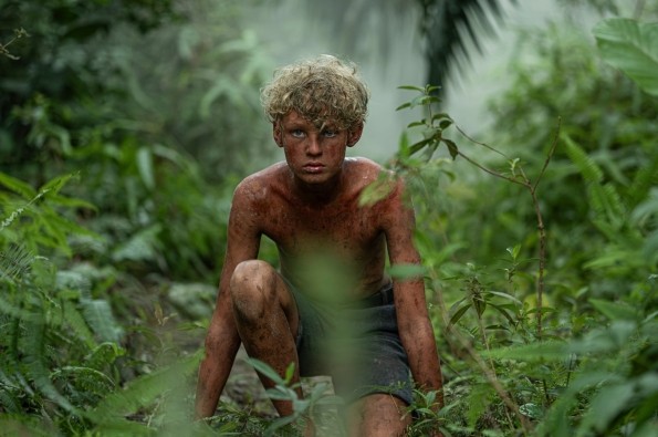 A person with curly blond hair sits on the ground in a dense jungle environment. The individual appears muddy and scratched, surrounded by lush green foliage and mist, evoking a wild and untamed setting.