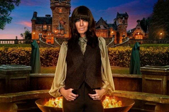 Promotional image from the fourth series of the British reality competition series The Traitors, showing host Claudia Winkleman standing in front of a large stone firepit with flames. Behind her is a grand castle surrounded by gardens, with two cloaked figures in the background. The scene is set at dusk with dramatic lighting.