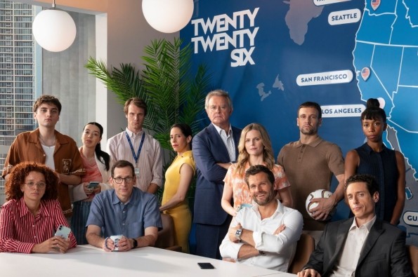 A group of people gathered around a modern conference table in a bright office. Behind them is a large blue wall map with city names like Seattle, San Francisco, Los Angeles, and Guadalajara, along with the text “TWENTY TWENTY SIX.” Two spherical pendant lights hang from the ceiling.