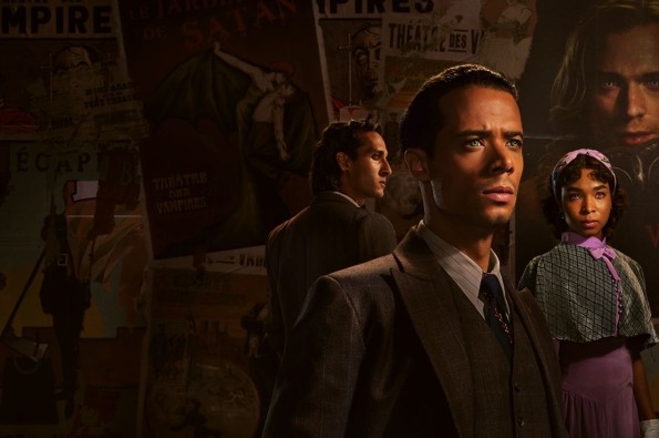 Three people dressed in vintage-style clothing stand in a dimly lit room with walls covered in old theatrical posters featuring words like “Vampire” and “Satan.” The figures wear formal suits and period dresses, evoking a dark, gothic atmosphere.