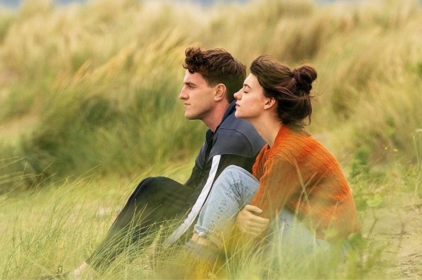 Two people sit close together on grassy sand dunes, dressed in casual clothing. One wears a dark jacket with striped sleeves, and the other wears a burnt orange jumper and light blue jeans. The setting is a natural coastal landscape with tall grass and soft, muted colours, suggesting a calm, outdoor scene.