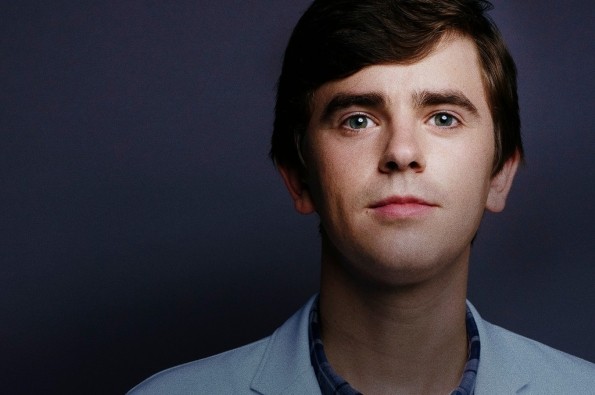 A person wearing a light blue medical-style jacket and a checked shirt standing against a dark, neutral studio backdrop, creating a clean and professional portrait composition.