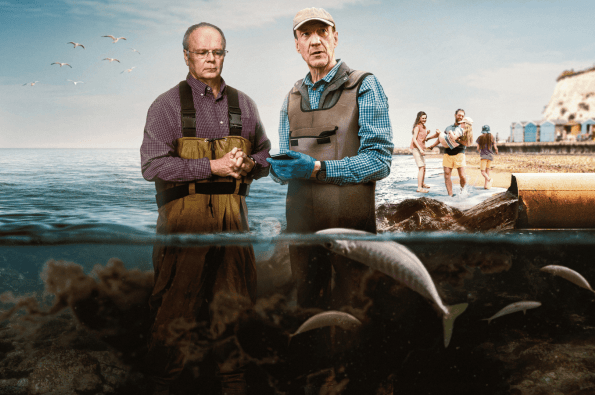 Two individuals wearing waders stand near the shoreline discussing marine conditions, with fish swimming underwater in the foreground and a coastal scene with beach huts, cliffs, and people near the water in the background.