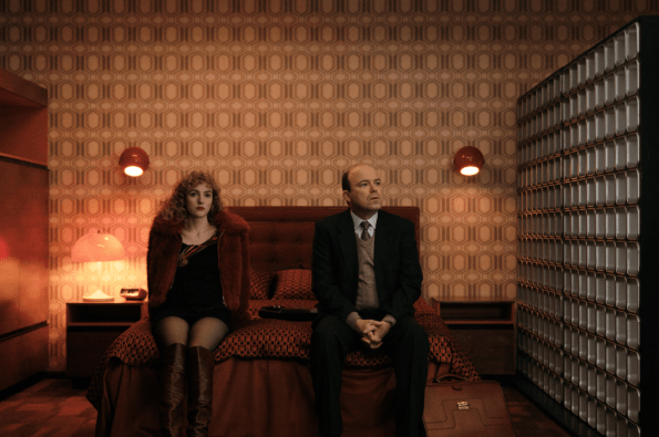Two people sitting on a bed in a dimly lit vintage-style room with patterned wallpaper, red lamps, and retro décor