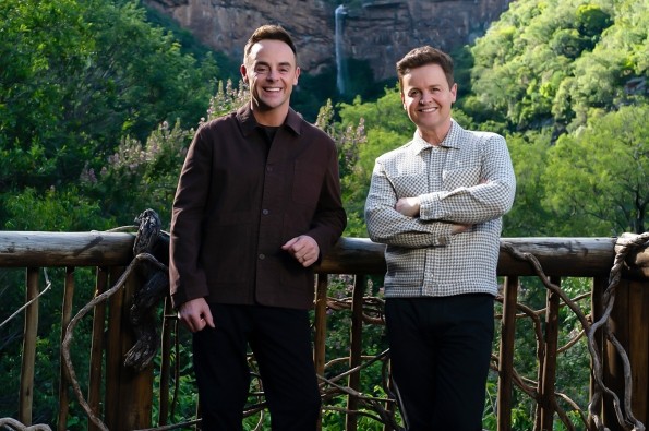 Two people standing on a wooden viewing deck surrounded by lush green forest, with a rocky cliff and waterfall in the background.
