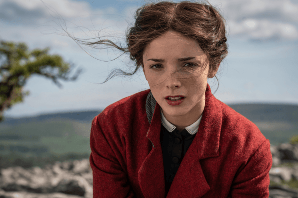 A person wearing a red coat sits outdoors on a rocky landscape with rolling hills and a partly cloudy sky in the background. Loose strands of hair blow in the wind. 