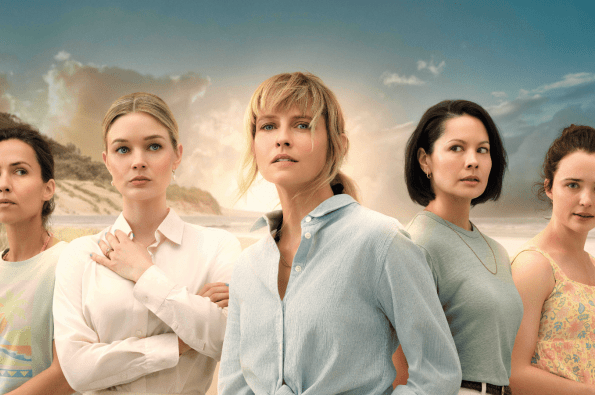 Five women wearing pale clothing stood looking suspiciously in different directions with a beach in the background