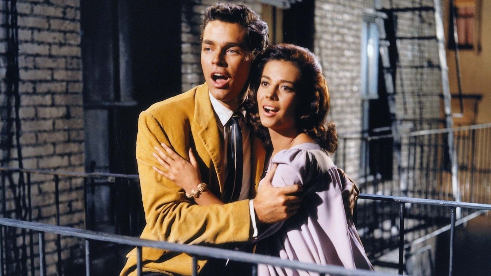 West Side Story key art- a man and a women stood by a fire escape is 1960s attire