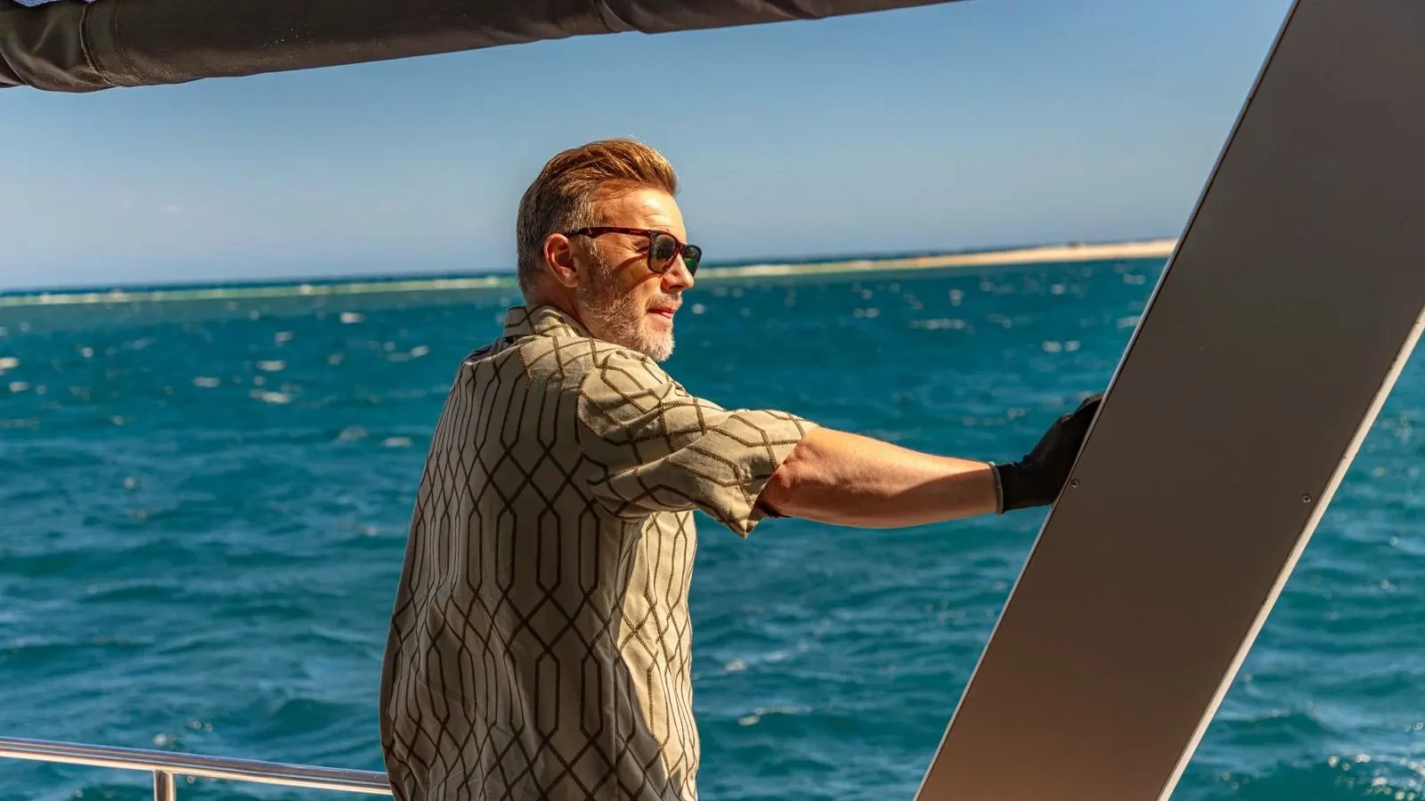 Gary Barlow stood on a boat with his right arm holding the side looking out at the sea