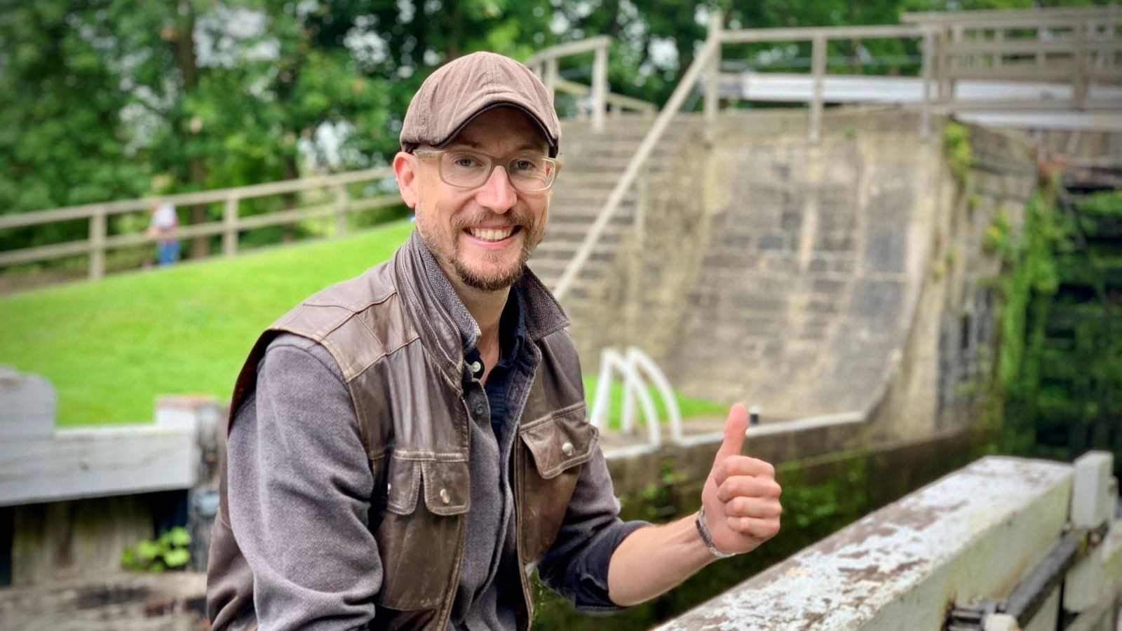 A man with a hat sat on the side of a canal with his thumb up