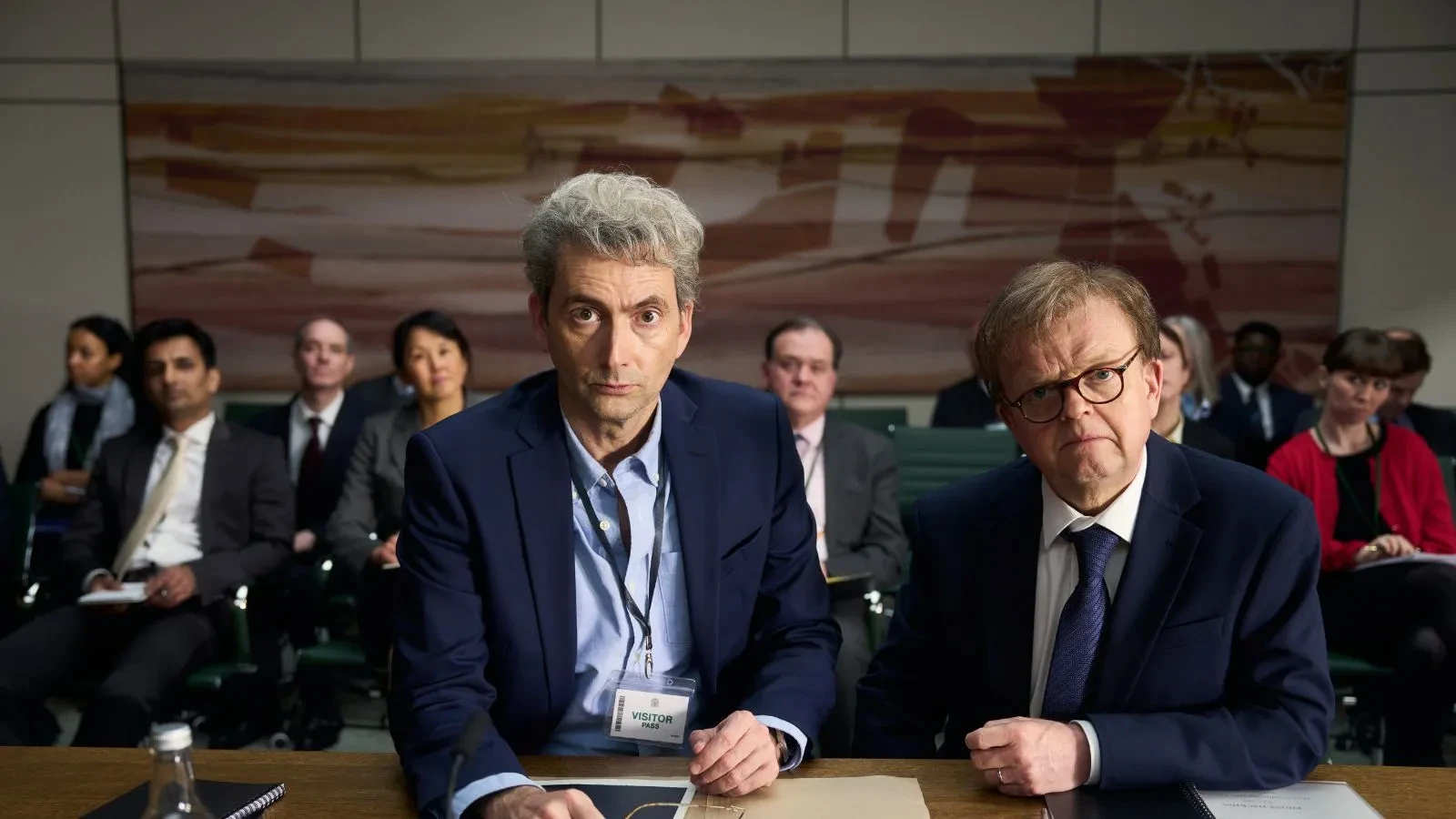 Two men sat a desk with a folder n front of them, dressed smart and others sat in chairs behind them