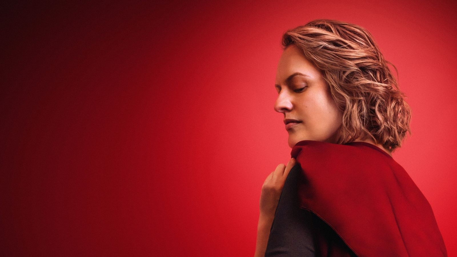 Elizsbeth Moss as June in a Handmaids tale, wearing a red cape against a red black ground