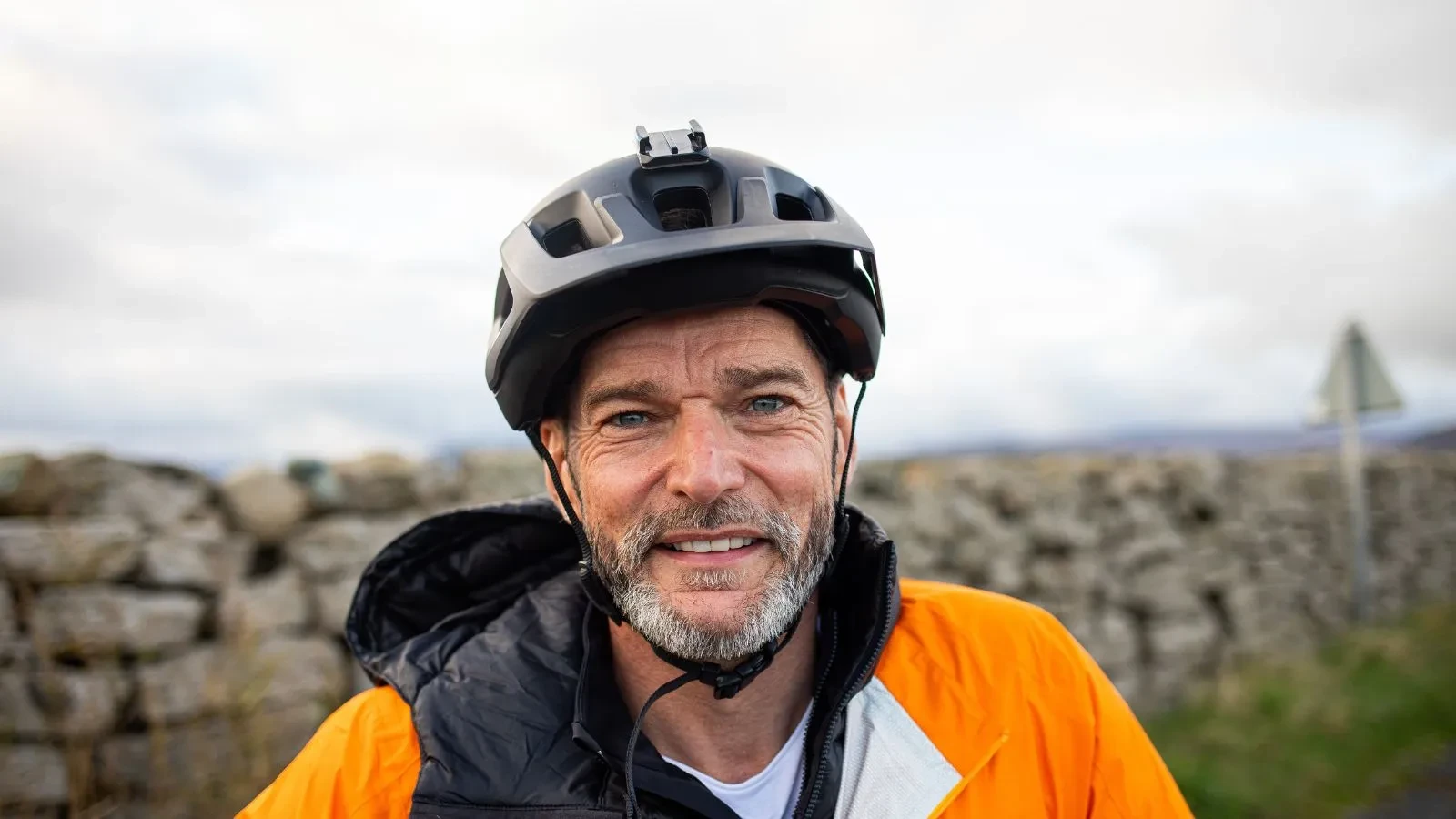 Fred Sirieix with a cycling helmet a bright orange jacket in front of an old wall