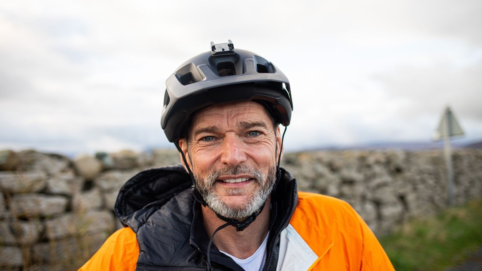 Fred Sirieix with a cycling helmet a bright orange jacket in front of an old wall