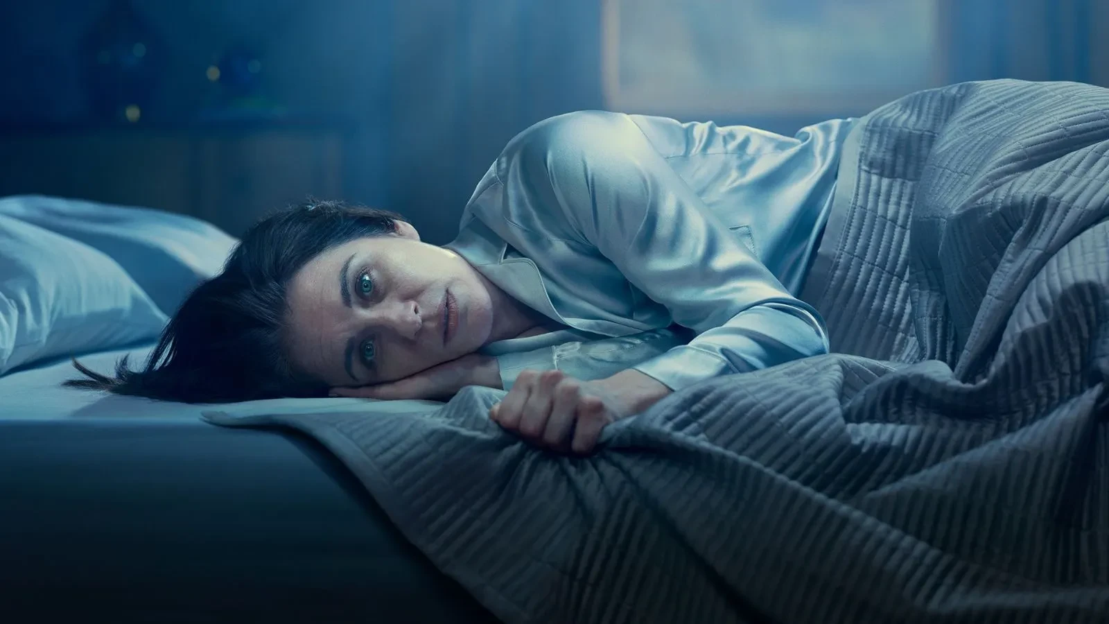 Woman with brown hair in silk pyjamas lying in bed staring at the camera