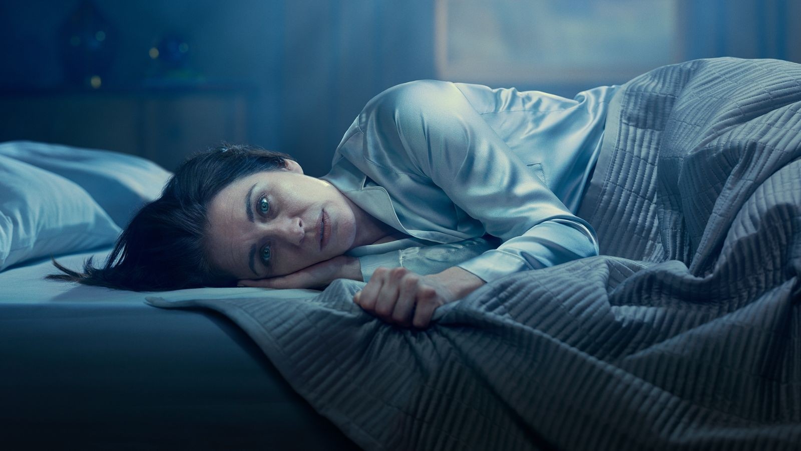 Woman with brown hair in silk pyjamas lying in bed staring at the camera
