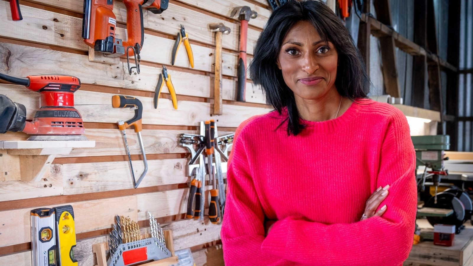 Women in bright pink jumper with her arms crossed and many tools on display behind her