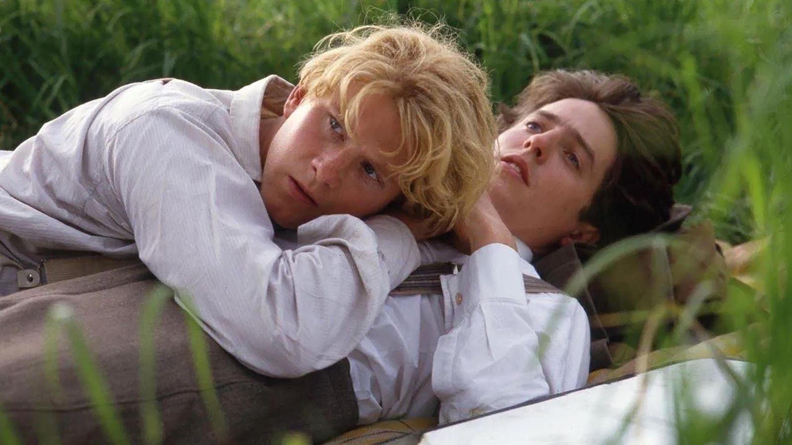 A blonde young man laying on a young man with brown hair in a green field both wearing shirts