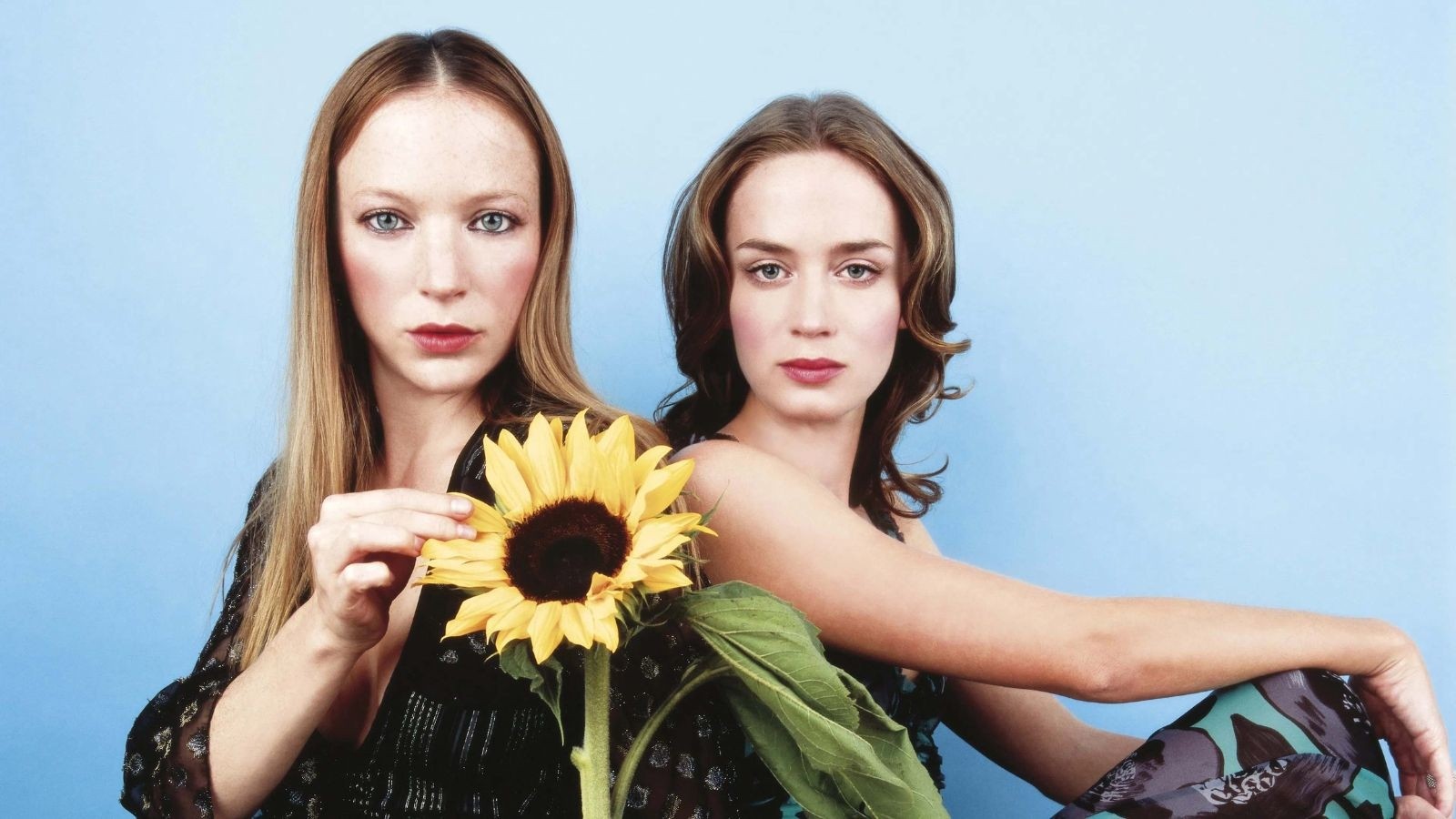 A blonde women holding a sunflower looking into the camera, a brunette women at next to her staring