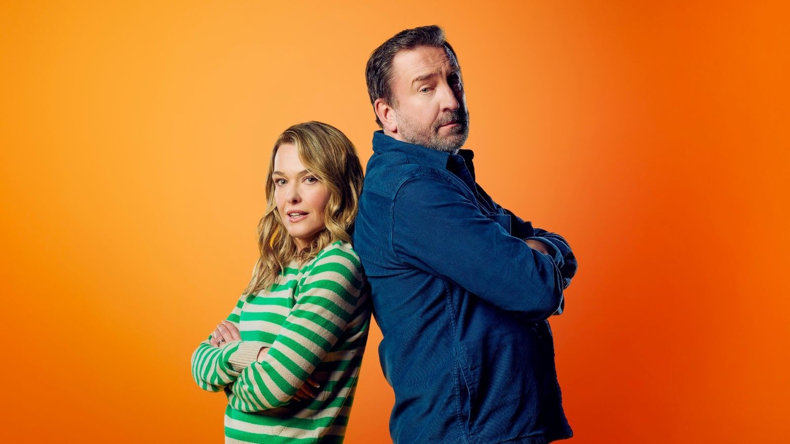 Women in green stripy jumper with her arms crossed back to back with a man in a blue shirt against an orange background
